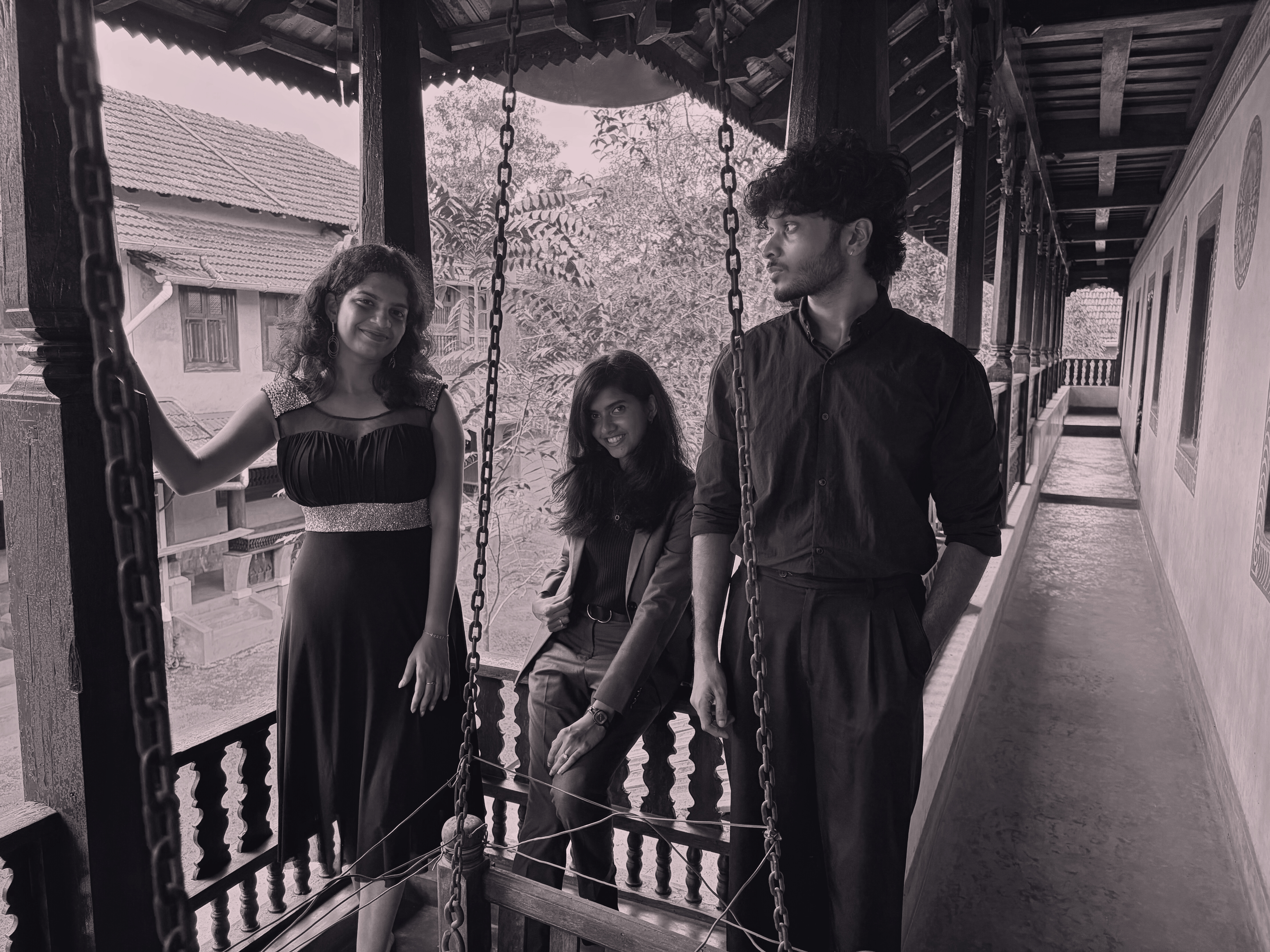 Black and white photo of three young and smart Indian individuals on a balcony of an Indian traditional house with traditional building and trees in the background.